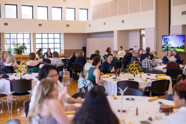 Guests seated at round tables during a banquet event in a bright, spacious hall. The tables are decorated with yellow napkins and small centerpieces, while some people chat, take photos, or look at their phones.